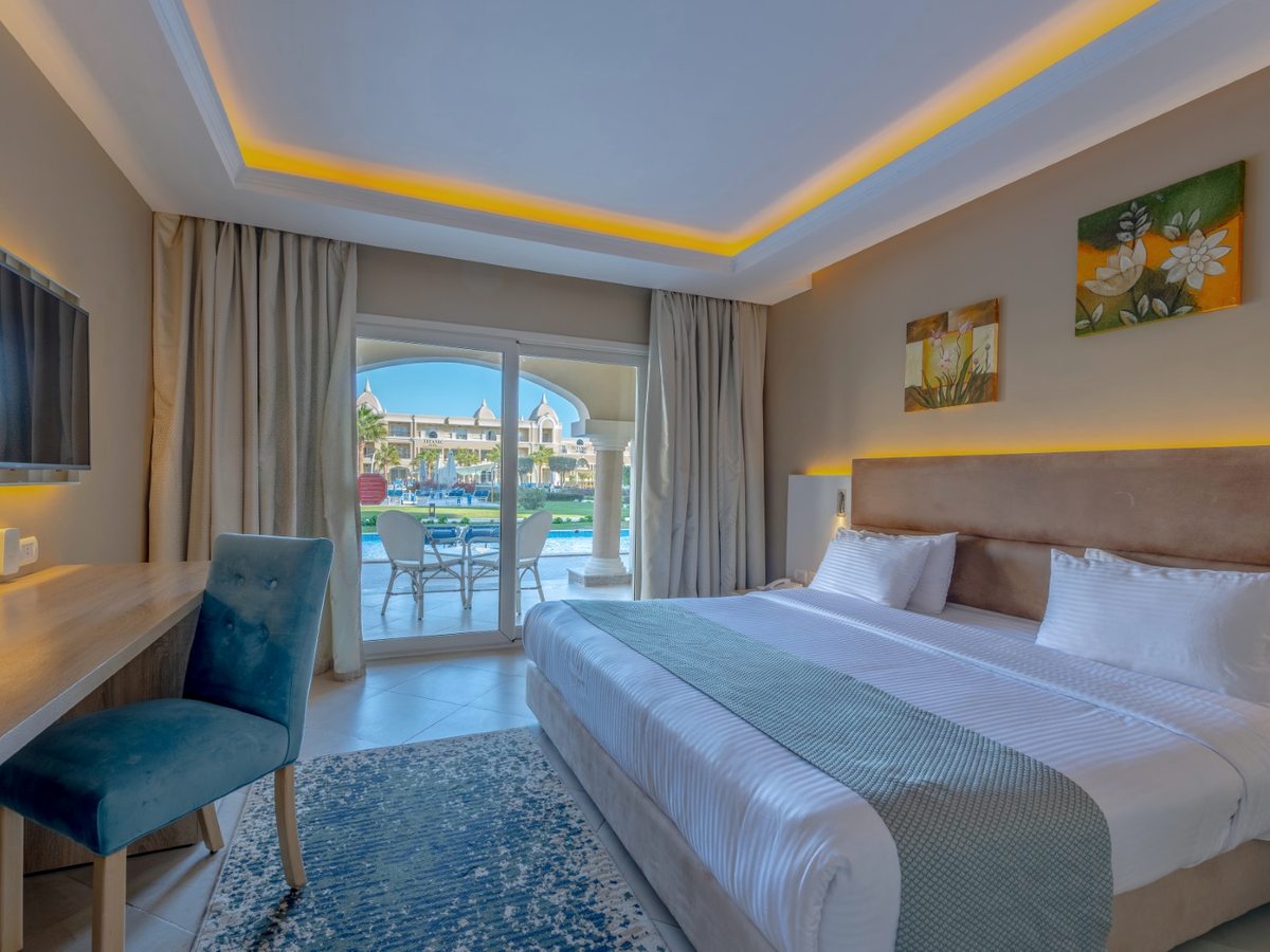 A bedroom with a double bed, blue and grey furnishings, a desk and a blue chair, and glazed doors opening onto a ground-floor terrace facing the pools and gardens.