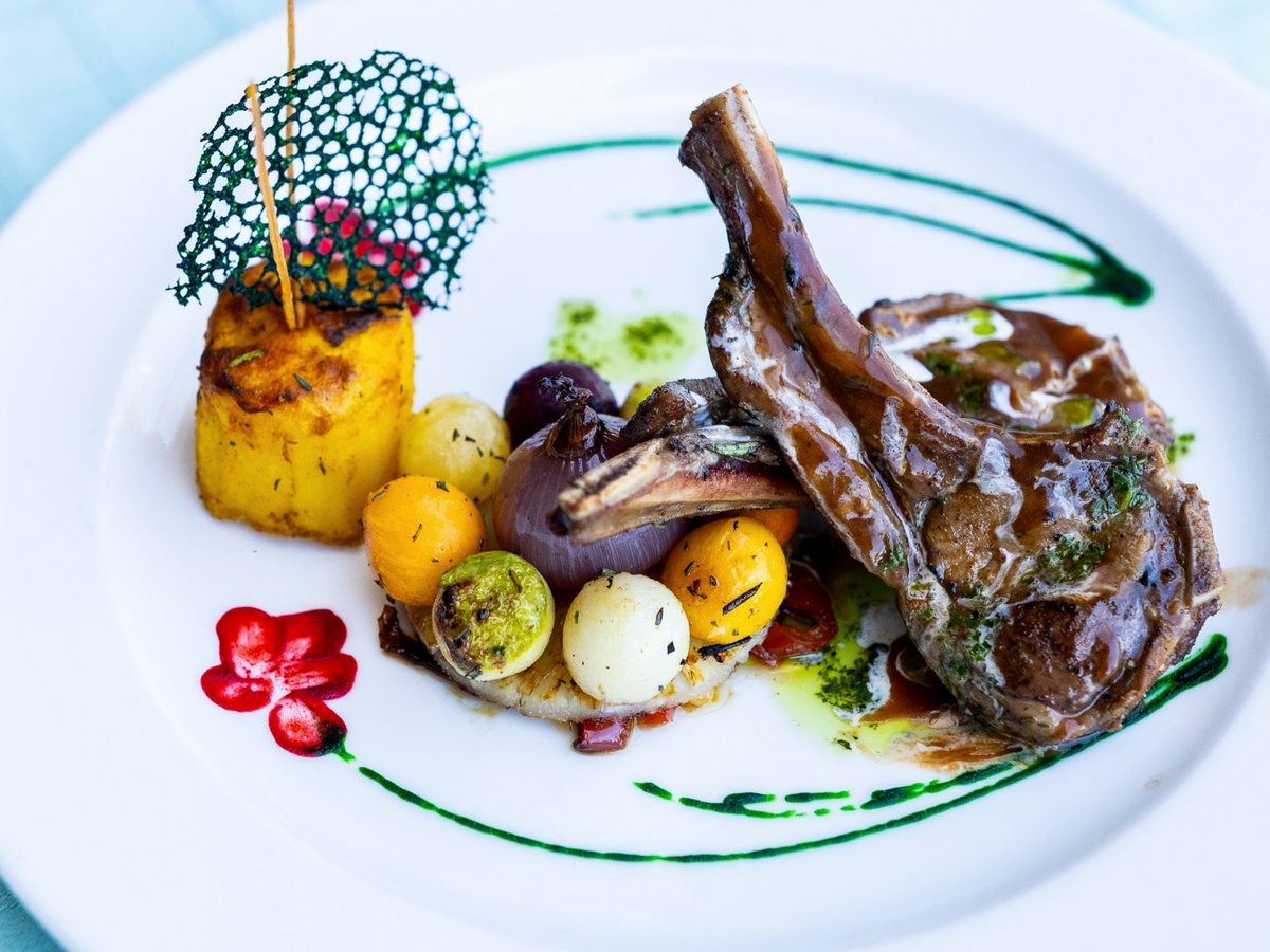 A white plate with a lamb chop, roast potatoes, baby vegetables and a dark sauce, finished with a green herb swirl.