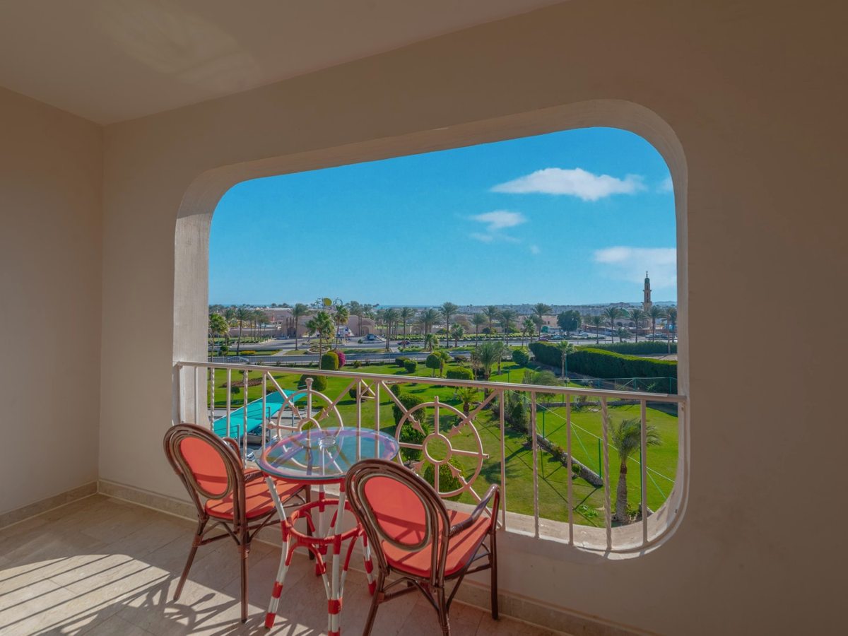 A room balcony with a table and two orange chairs behind a white railing, looking over the resort gardens and the road beyond.