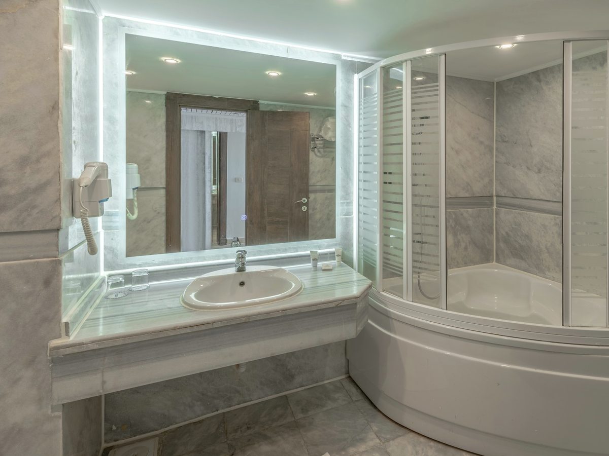 A bathroom with a marble-topped vanity, a large mirror, a hairdryer and a deep corner bath enclosed by curved sliding glass panels.