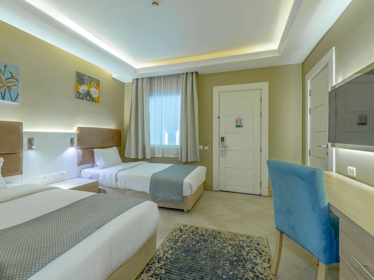 A bedroom with two single beds, tan padded headboards on white panels with reading lights over them, a desk, a wall-mounted television and a curtained window.