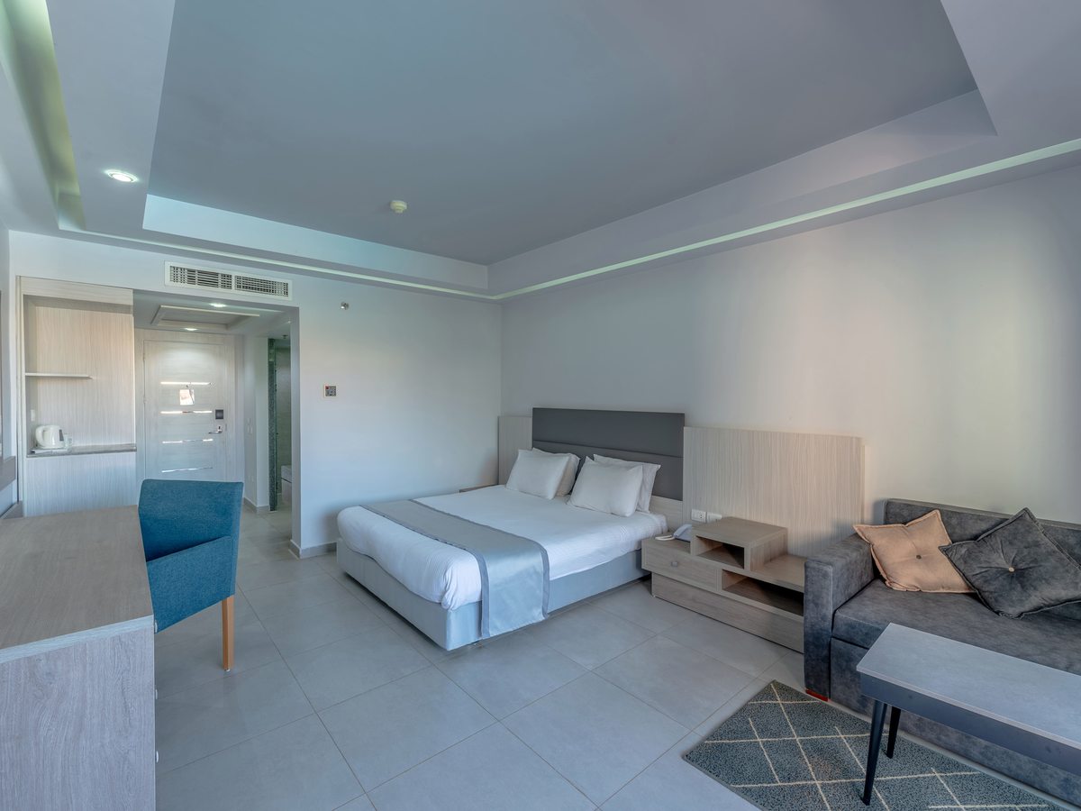 A spacious room with a double bed, a grey sofa and low table on a patterned rug, a desk with a blue chair and a tea tray by the entrance hall.
