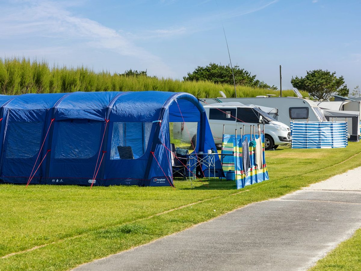 A large blue family tent pitched on mown grass beside a tarmac lane, with touring caravans and a striped windbreak behind.