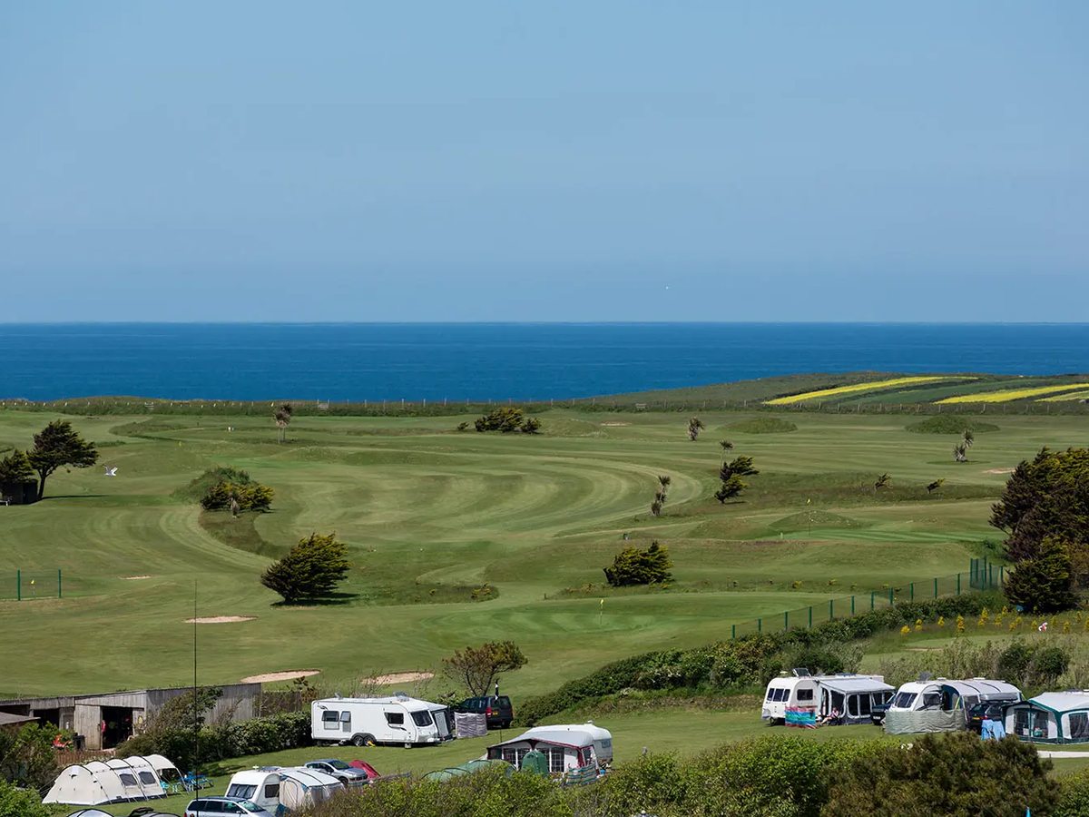 A wide green golf course dotted with bunkers and wind-bent trees running down to a deep blue sea, with tents and touring caravans in the foreground.