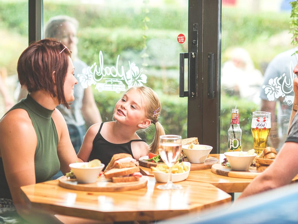 A woman and a young girl at a wooden table with sandwiches and chips, beside a glass door carrying the Woody's logo.