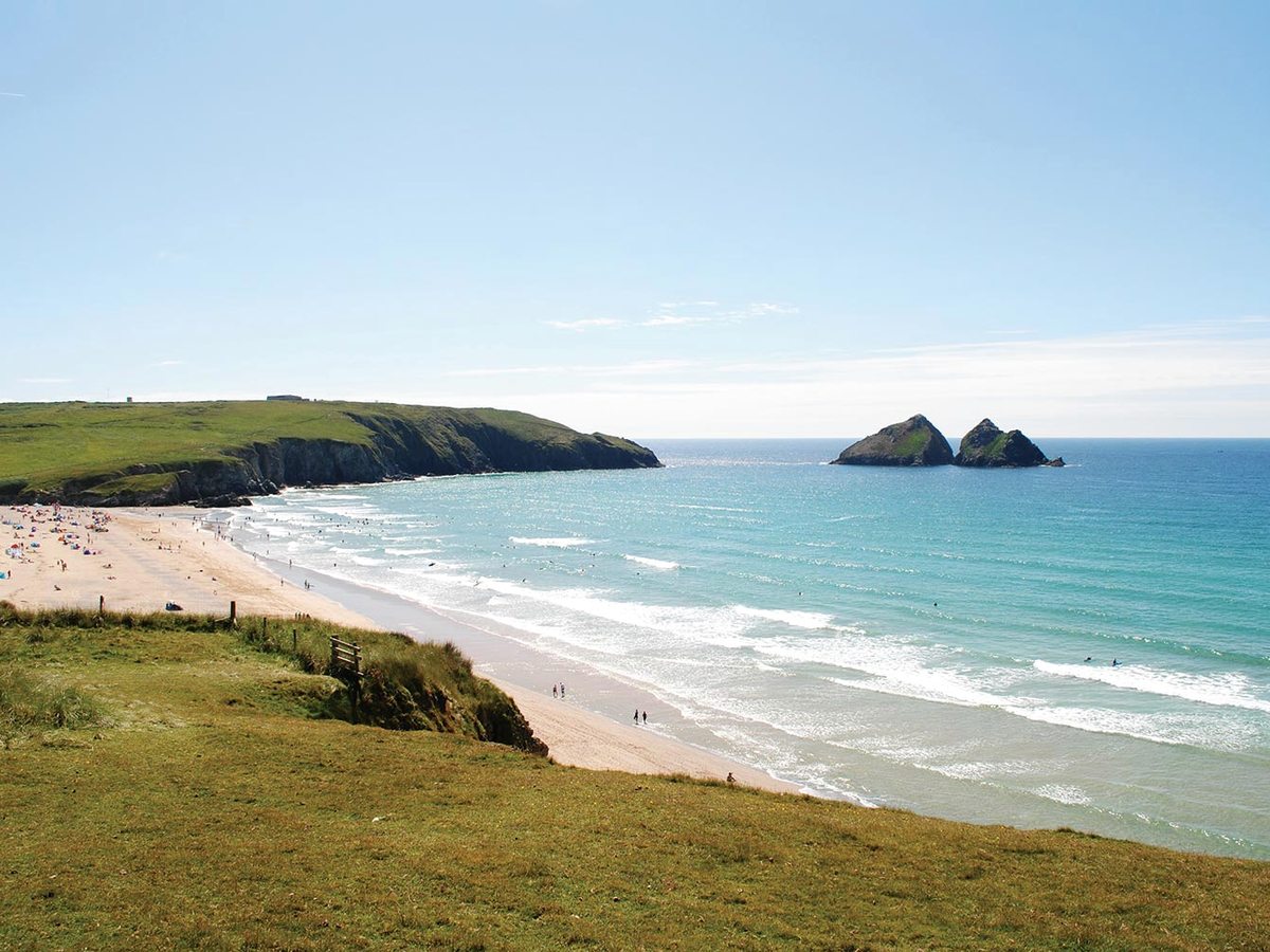 A long sandy beach curving between grassy headlands with turquoise surf, and two rock stacks standing offshore.