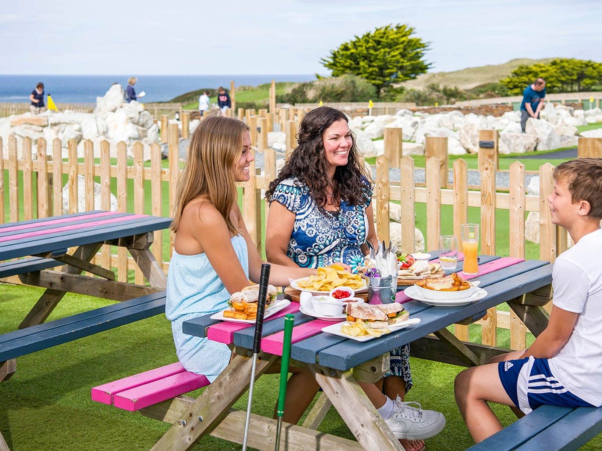 A family eating sandwiches and chips at a pink and blue picnic table beside the mini golf course, with the sea behind.