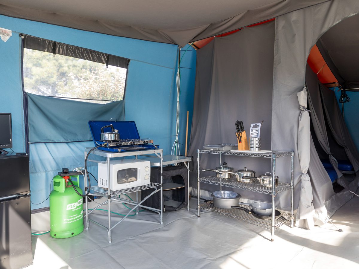 The inside of a canvas tent with a two-ring gas stove, a microwave, a fridge and a shelf of pans, with a bedroom compartment through the doorway.