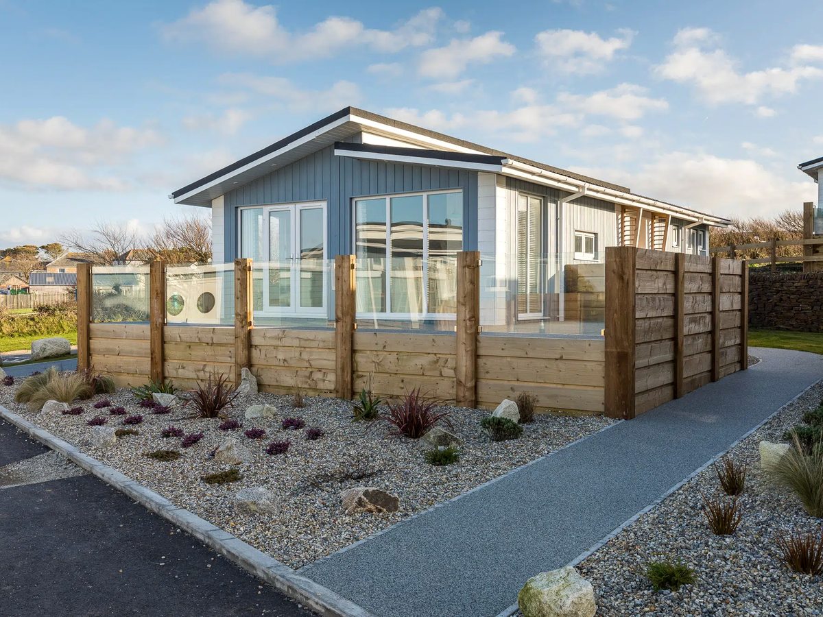 A blue and white timber-clad lodge with large windows and a glass-topped fence, set in gravel landscaping.