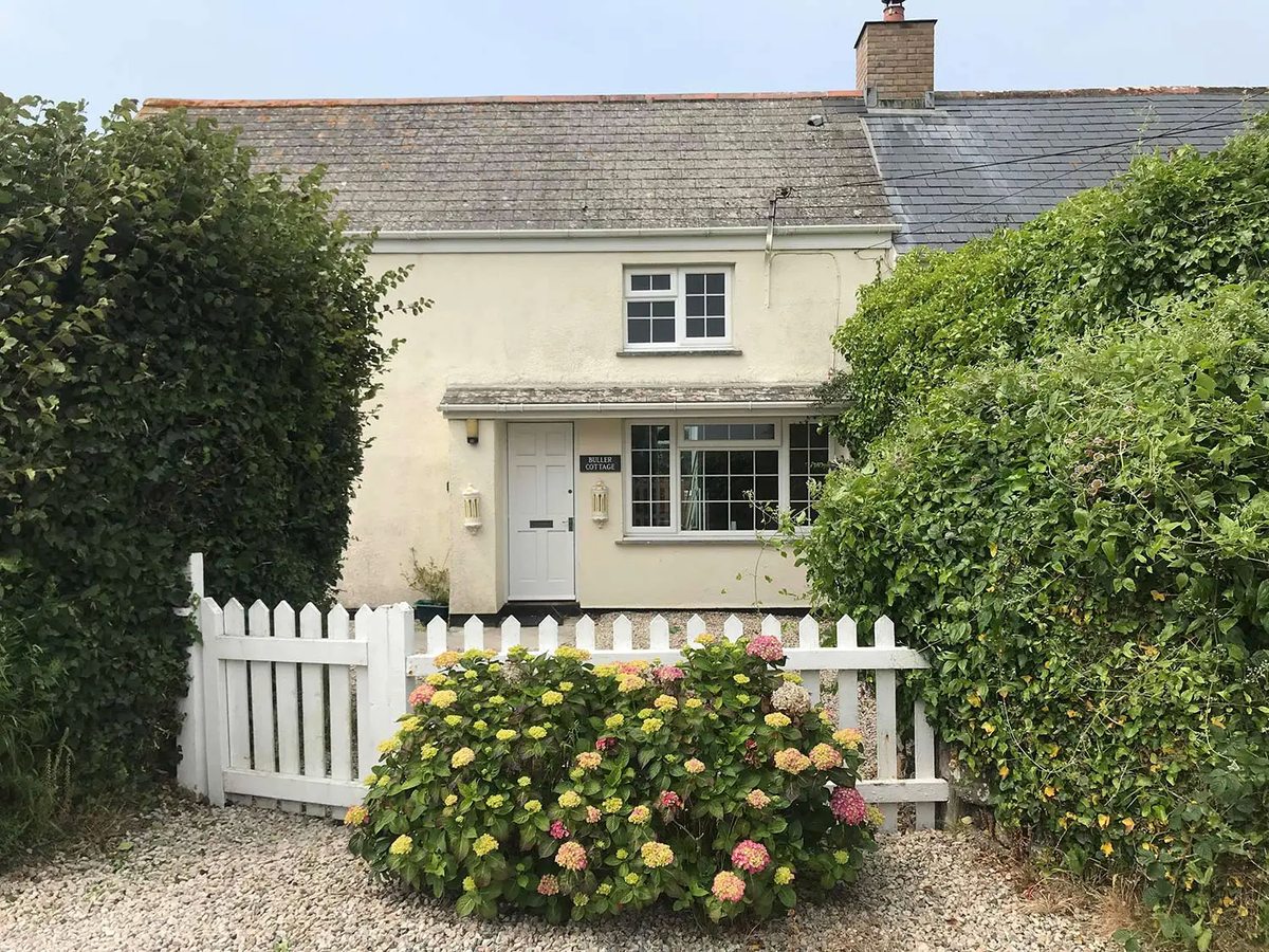 A cream rendered cottage with a slate roof, a white picket gate and hydrangeas in the front garden.