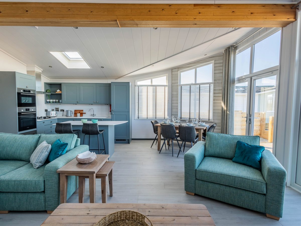 An open-plan lodge living room with teal sofas, a blue kitchen with an island and a dining table by full-height windows.