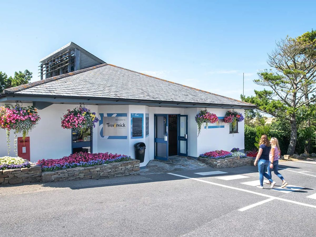 The Trevornick reception building, a low white building with hanging flower baskets and a Welcome to Trevornick sign, with two people crossing the car park.