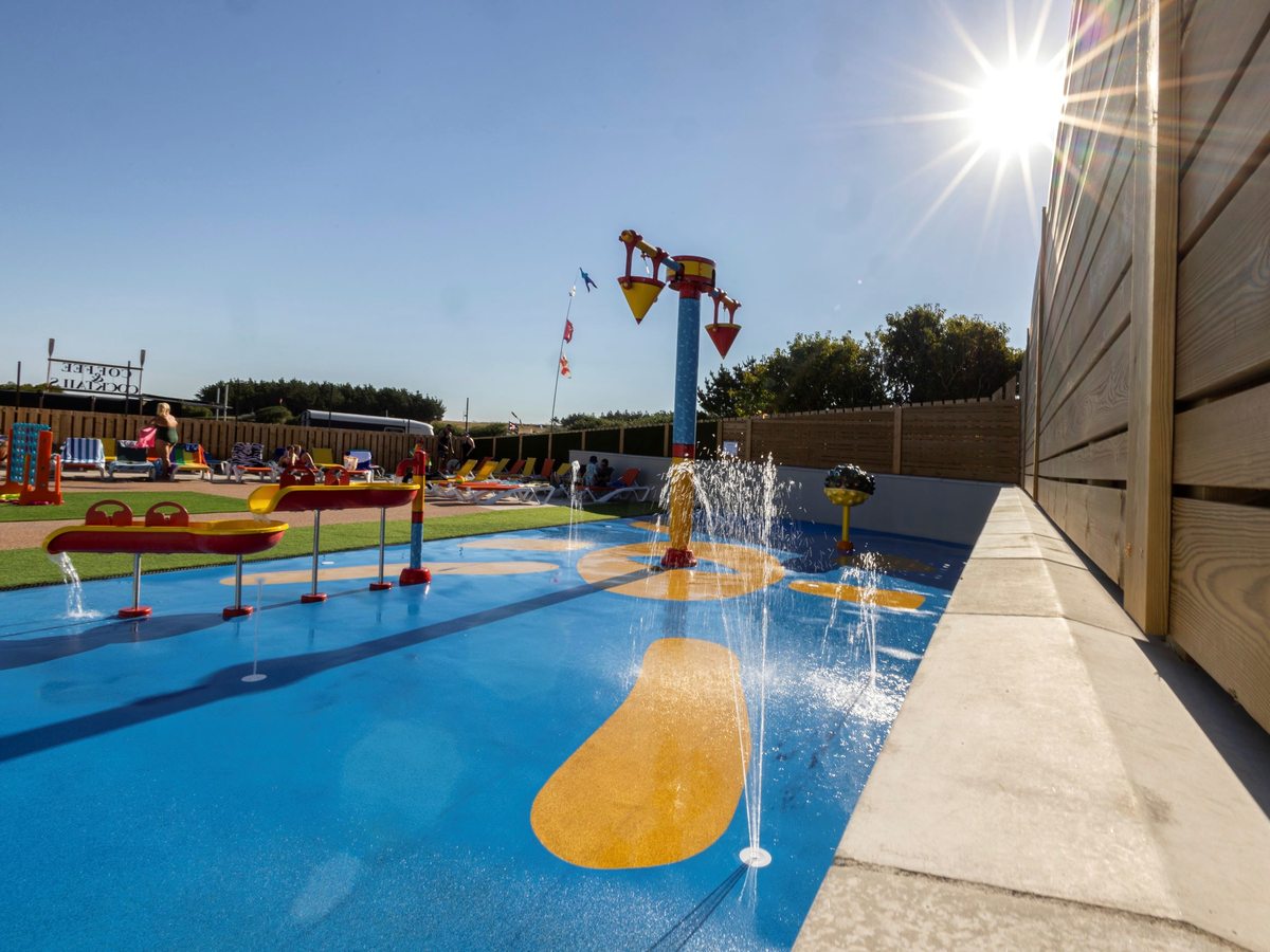 A blue and yellow splash pad with water jets and tipping buckets, with sun loungers on the terrace behind.