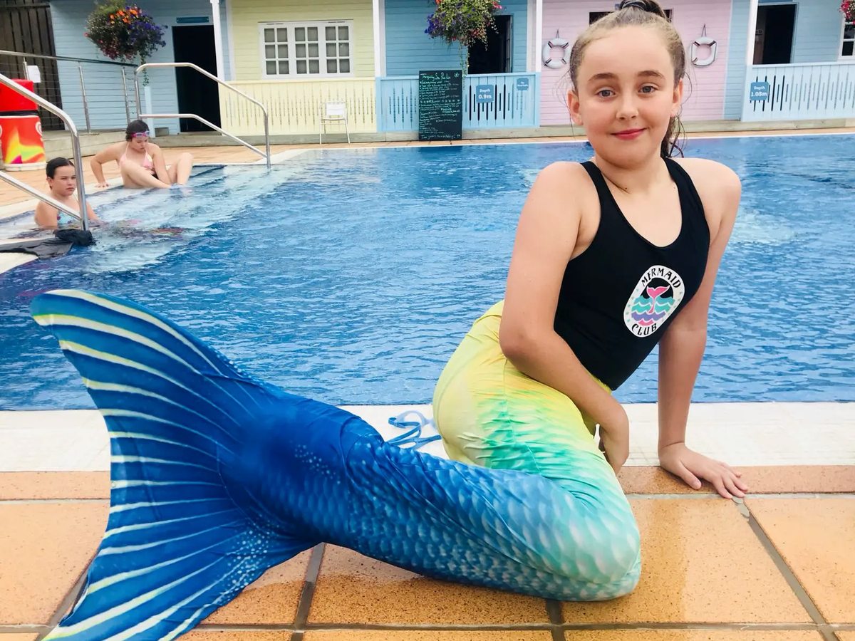 A girl in a blue and green mermaid tail sitting at the edge of the outdoor pool, with other children swimming behind.