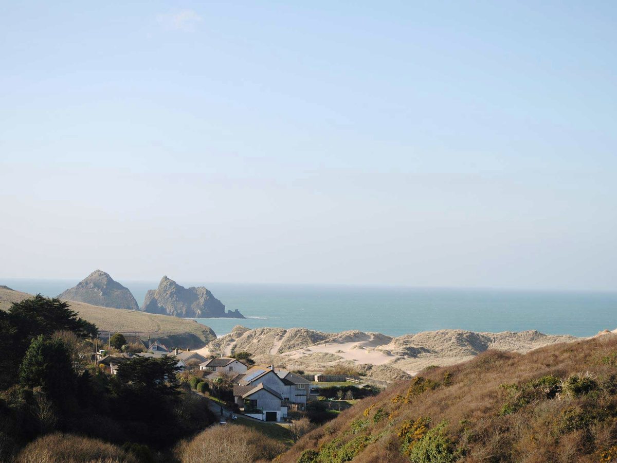 Houses and gardens on a hillside above sand dunes, with the sea and two rock stacks beyond.