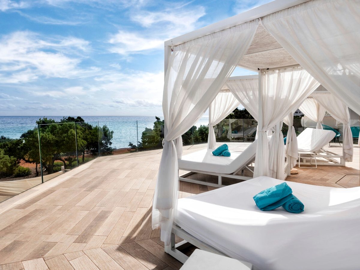 White four-poster daybeds with gauzy curtains and folded turquoise towels on a pale wooden deck, with a glass balustrade and the sea beyond.