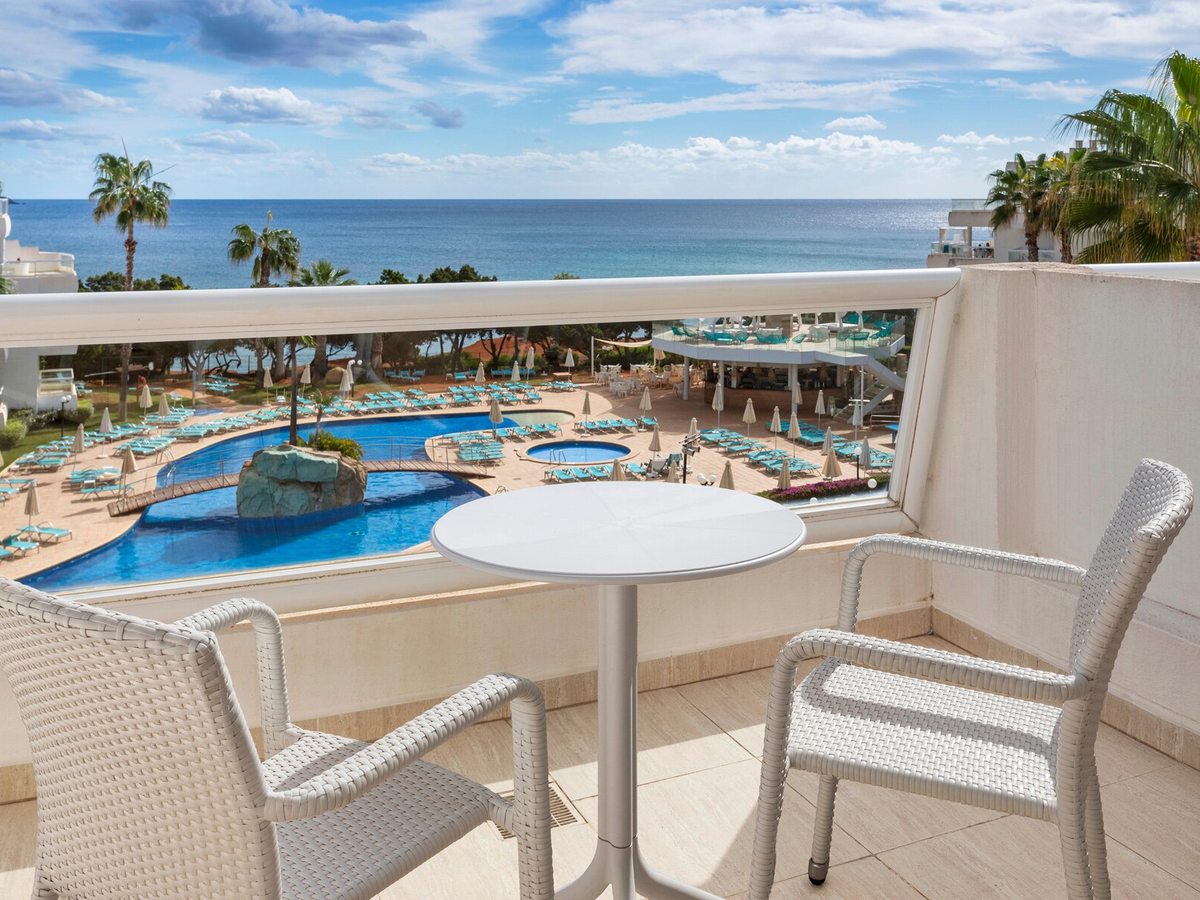 A round white table and two woven armchairs on a room balcony, with the resort's pools, palms and the sea below.