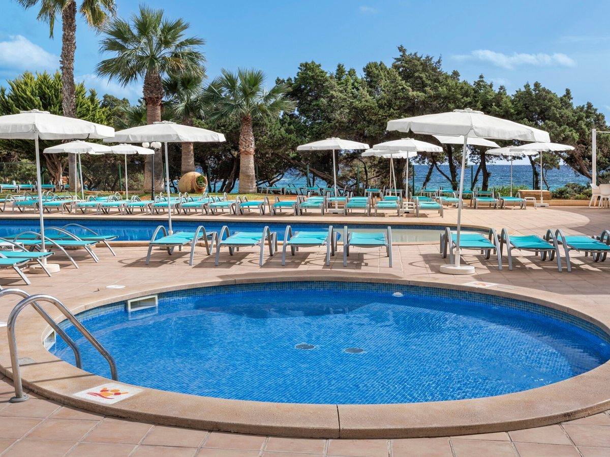 A circular blue-tiled pool with a metal handrail, ringed by turquoise sun loungers and white parasols, with pines and the sea behind.