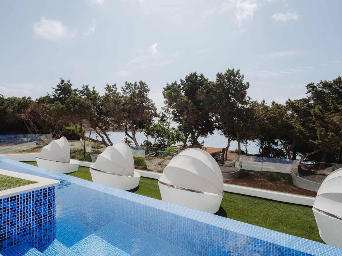 A blue mosaic-tiled pool with white shell-shaped daybeds on artificial grass beside it, and pines and the sea beyond a glass balustrade.