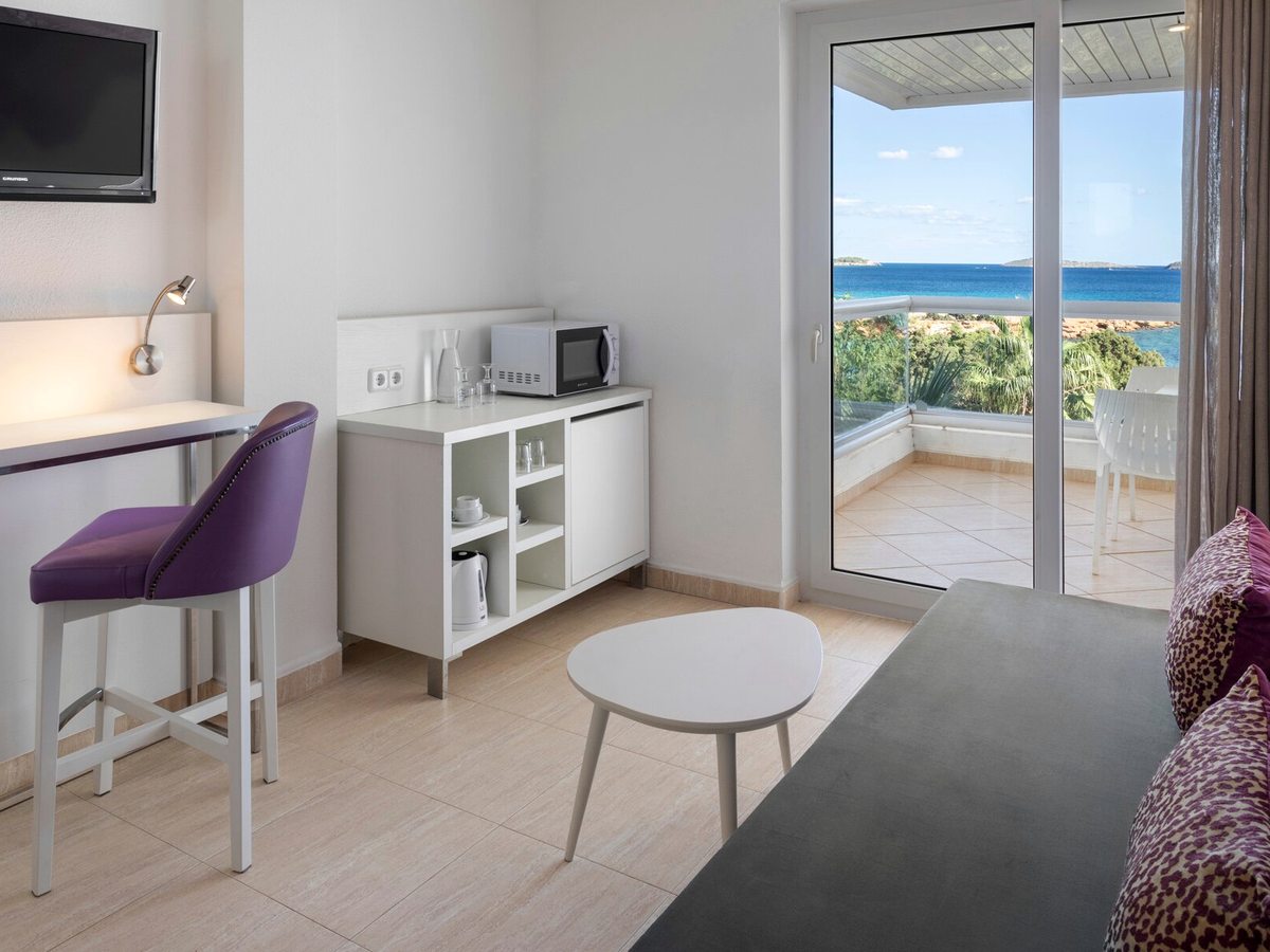 A room living area with a white unit holding a microwave, kettle and glasses, a counter with a purple stool, a sofa, and glass doors onto a terrace with a sea view.