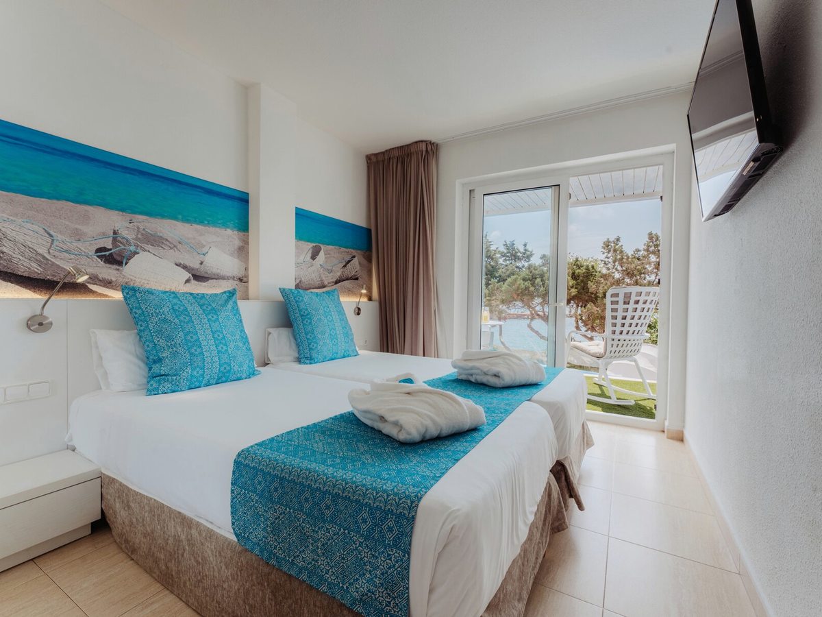 A room with two single beds pushed together under a beach-photograph headboard panel, a wall-mounted TV, and glass doors onto a balcony with a chair and the sea beyond.