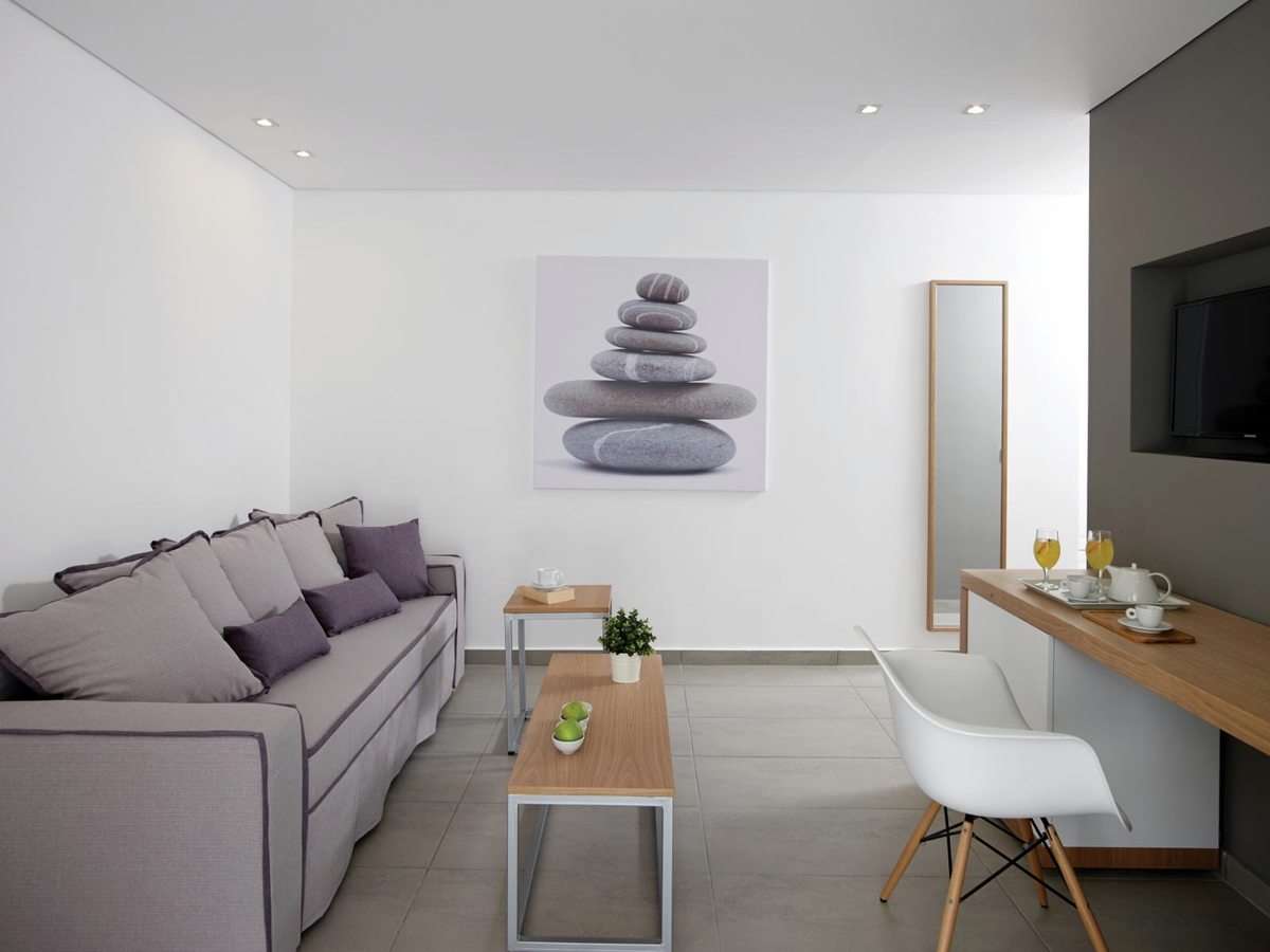 Suite sitting area with a long mauve sofa, a low coffee table, a wall-mounted television and a white desk chair.