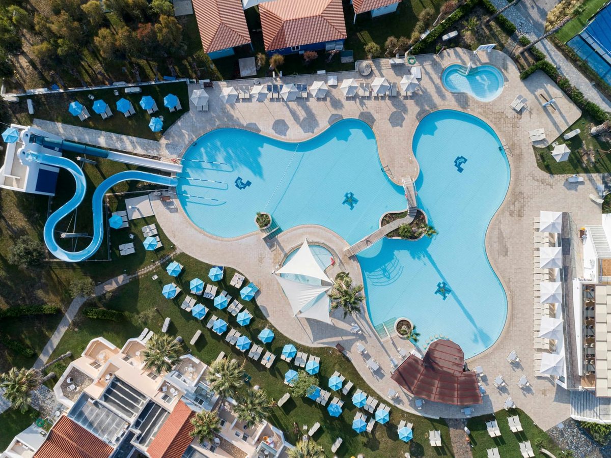 Overhead view of a freeform pool complex with a smaller round pool, rows of blue parasols and loungers, and two waterslides curving down at one side.