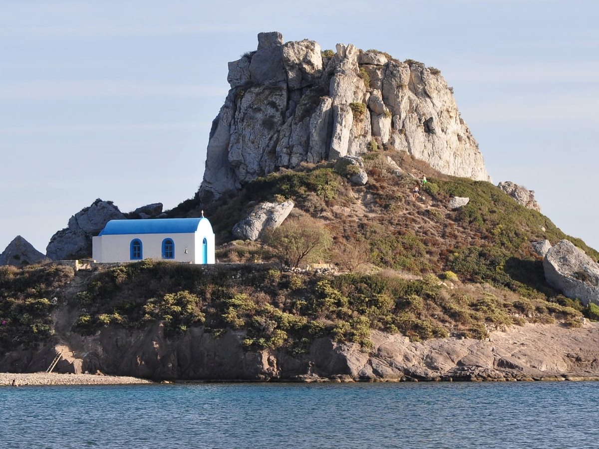 Small white chapel with a blue roof on a rocky islet rising from the sea, with scrub-covered slopes above.