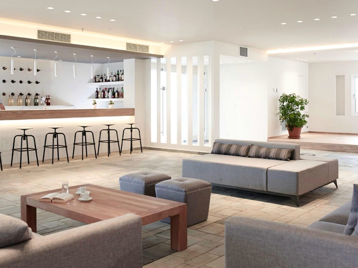 Indoor bar with a long counter and bar stools backed by bottle shelves, with grey sofas and low wooden tables in front.