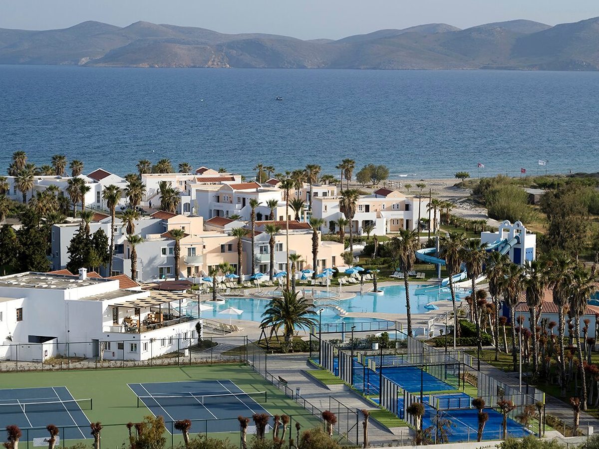 Elevated view of the resort: white and pastel blocks among palms, the pool and its waterslide behind, blue tennis and padel courts in the foreground and the sea and mountains beyond.