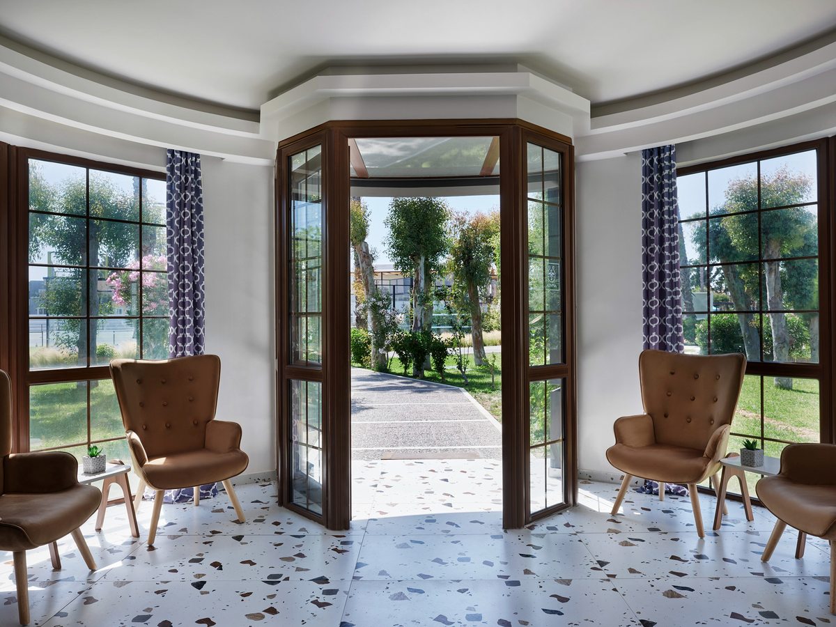 Bay-windowed lounge with tan buttoned armchairs and small side tables on a white terrazzo floor, with tall glazed doors open onto a garden path.