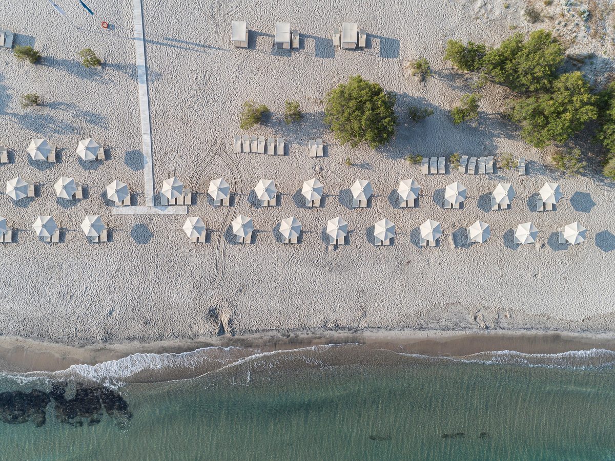 Overhead view of rows of cream parasols and sunbeds on pale sand, with clear shallow water breaking along the shore.