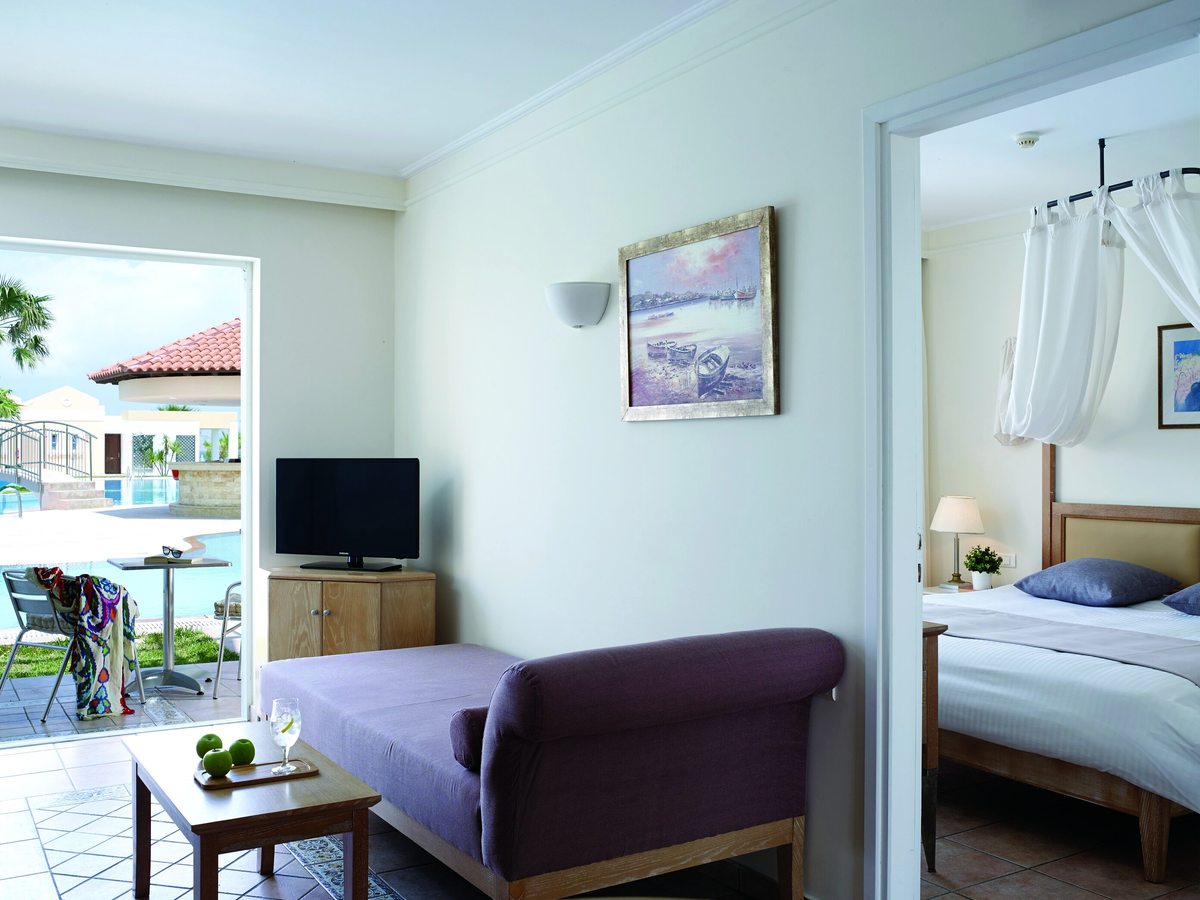 Family suite living area with a purple daybed, coffee table and television, patio doors onto a poolside terrace and a bedroom through an open doorway.