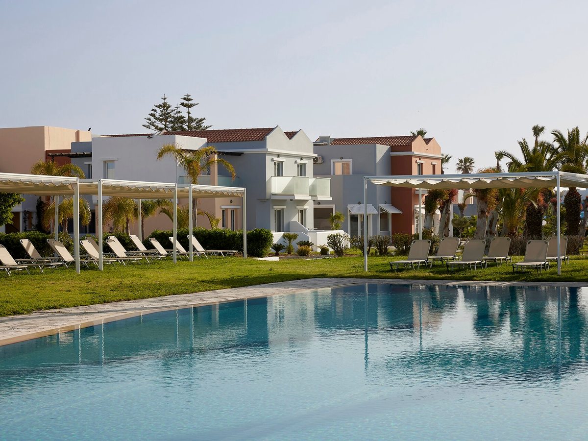 Long rectangular pool edged with lawn, sun loungers and white shade pergolas, with pastel two-storey blocks behind.
