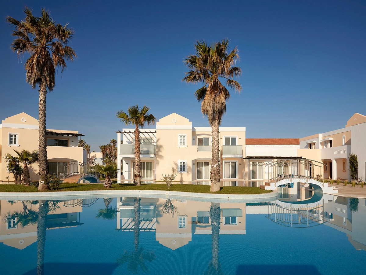 Still pool reflecting cream two-storey buildings and tall palms, with a metal footbridge crossing it.