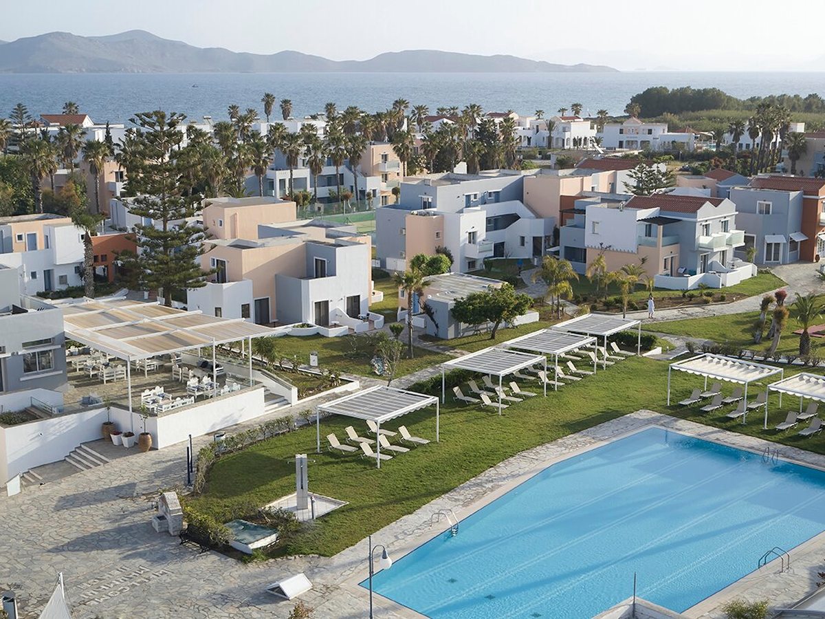Elevated view over the resort: cream and white low-rise blocks with red-tiled roofs among lawns, palms and paved paths, a rectangular pool in the foreground and the sea and hills behind.