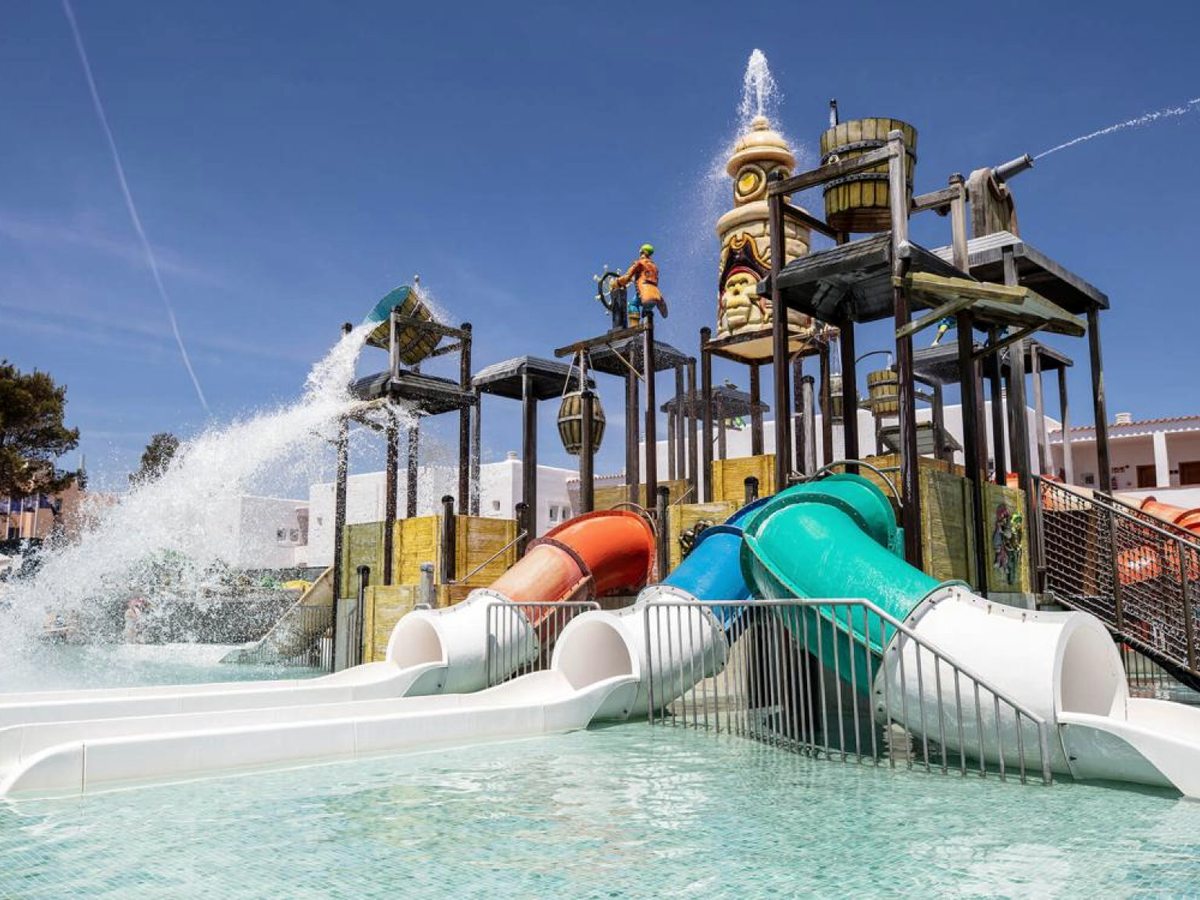 A timber pirate-ship water-play structure with tipping barrels and a cannon spraying water, and orange and green tube slides running down into a shallow pool.