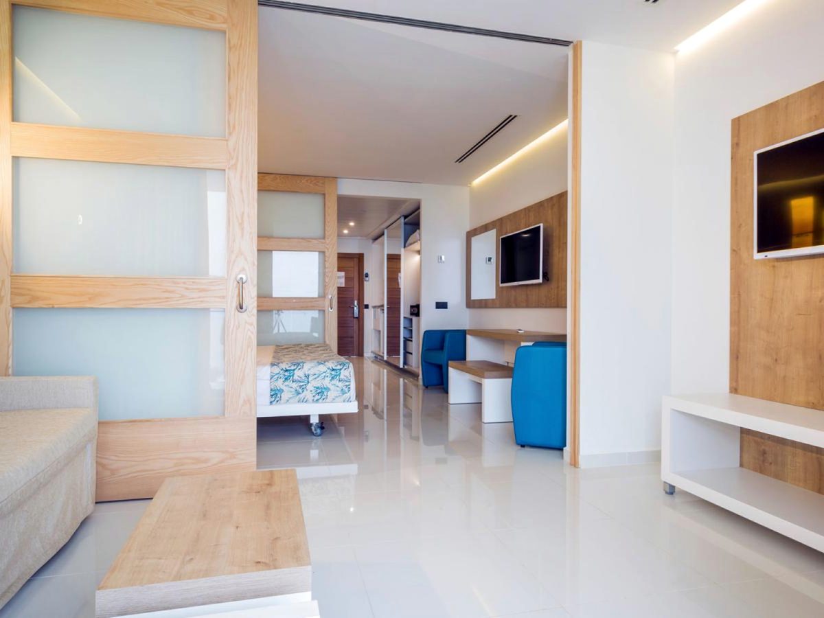 A suite with a sofa and low table in the foreground and a frosted sliding partition drawn back to a sleeping area, with a desk and television opposite.