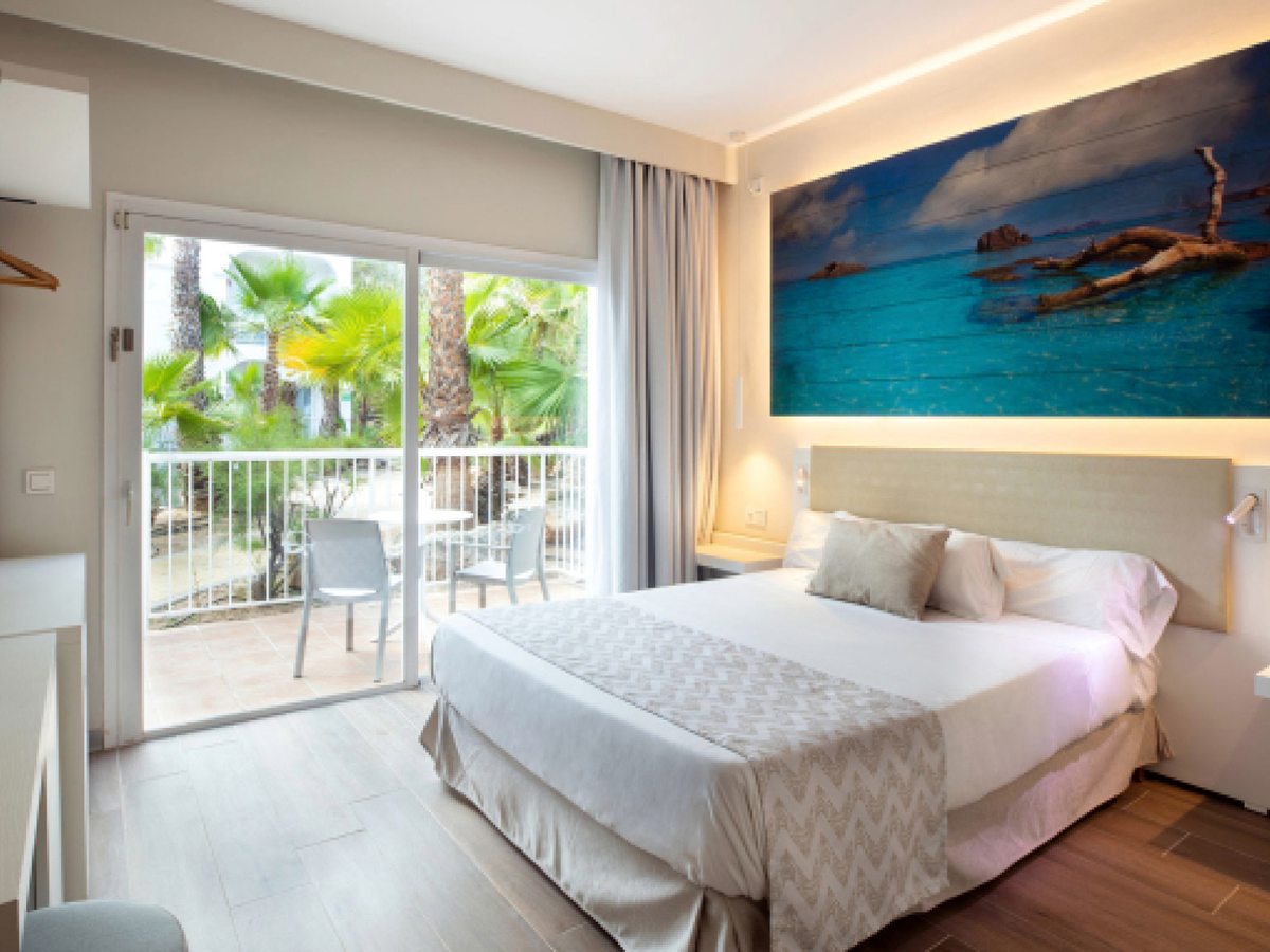 A double room with a wide bed, white bedding and bedside lamps, a large sea photograph above the bed and sliding doors to a balcony with palms beyond.