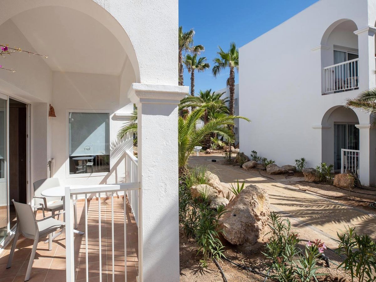White low-rise accommodation blocks with arched balconies and ground-floor terraces, set among palms, bougainvillea and gravel planting.