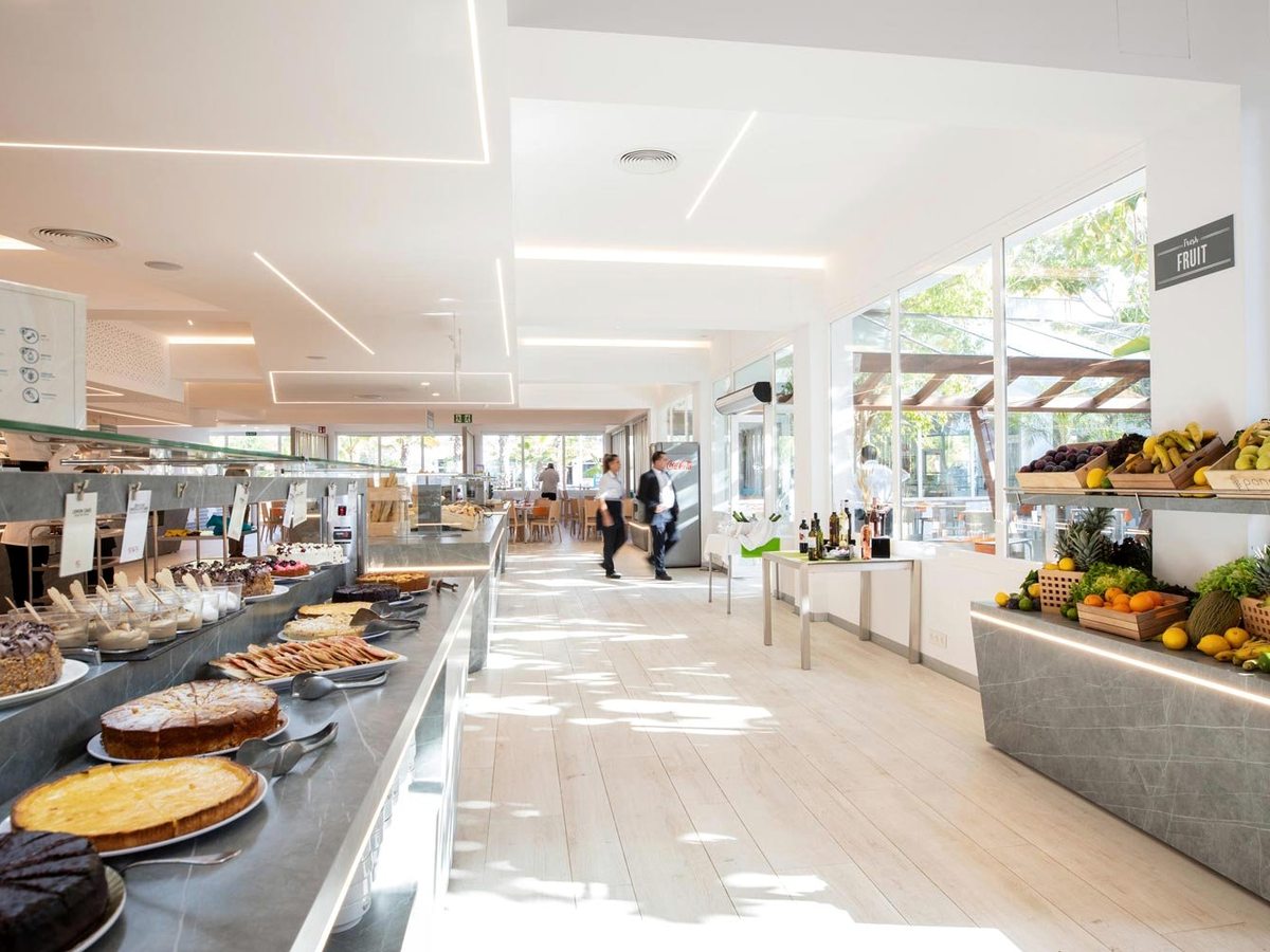A long buffet hall with food counters down one side, a fruit display and dining tables by full-height windows.