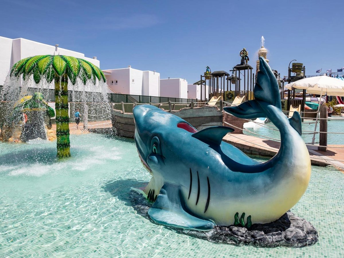 A shallow play pool with a large shark sculpture, a green palm-tree fountain raining water and the pirate structure behind.