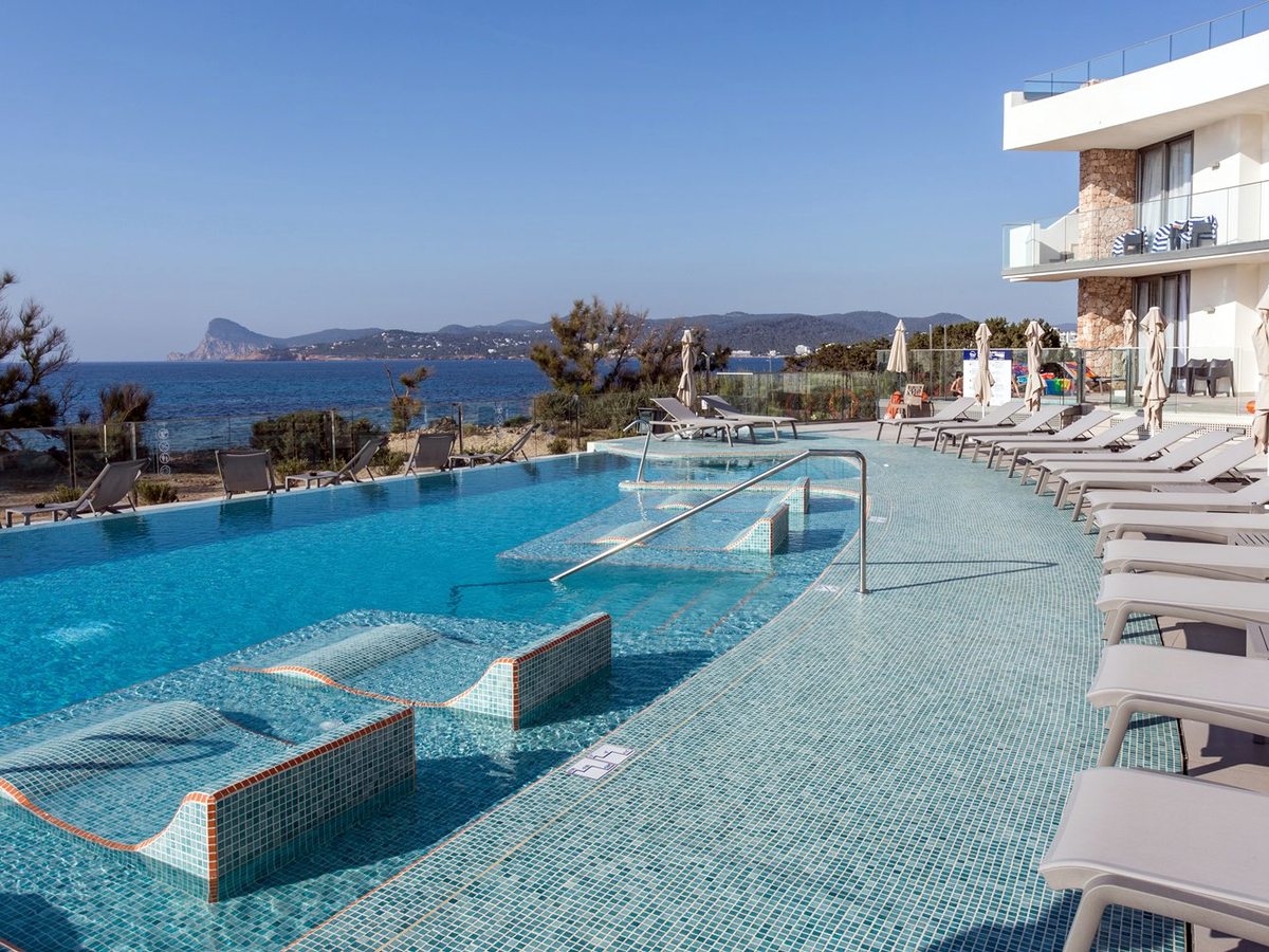 A long pool with tiled in-water loungers and steps, rows of sun loungers alongside and the sea and a headland beyond.