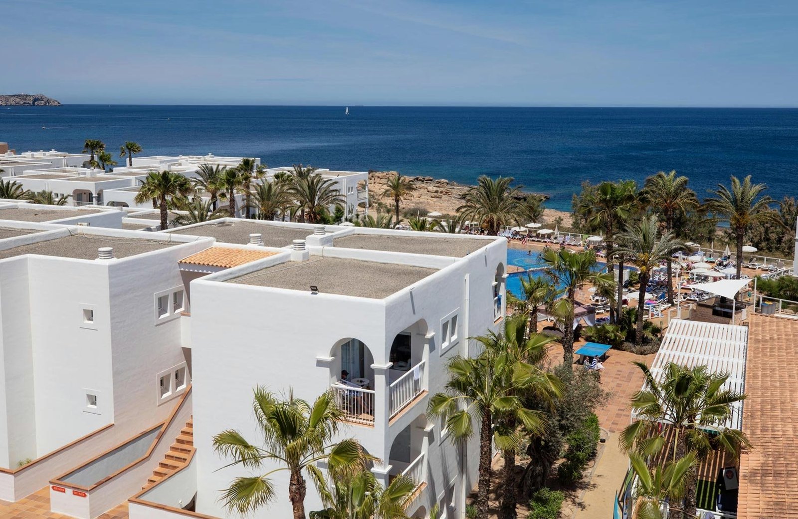 The white low-rise buildings of TUI BLUE Aura among palms above Port des Torrent, with the pool terrace, the shore and the sea beyond.