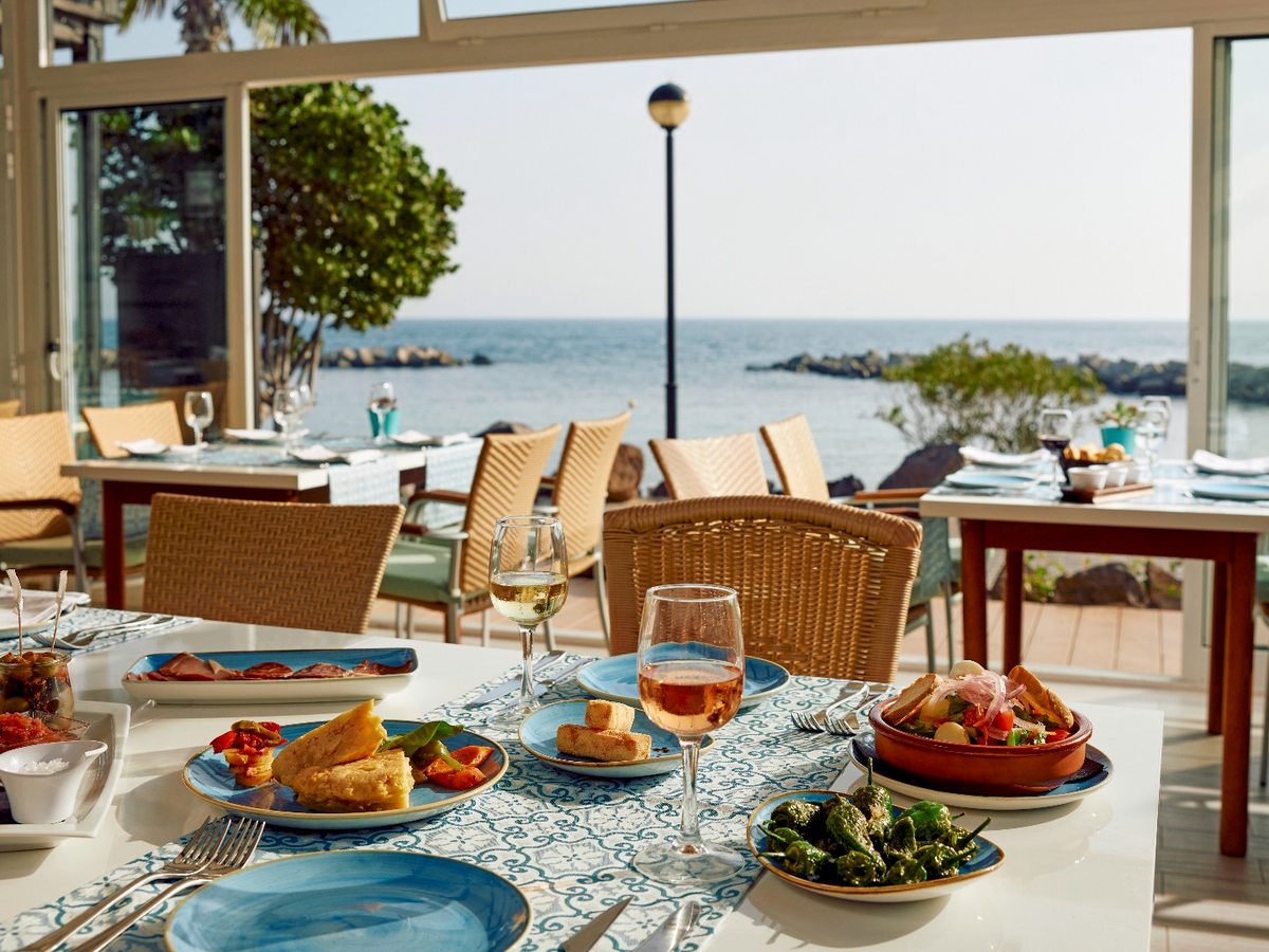 A restaurant terrace of laid tables and wicker chairs behind a glass windbreak, with plates of food in the foreground and the rocky shoreline and sea beyond.