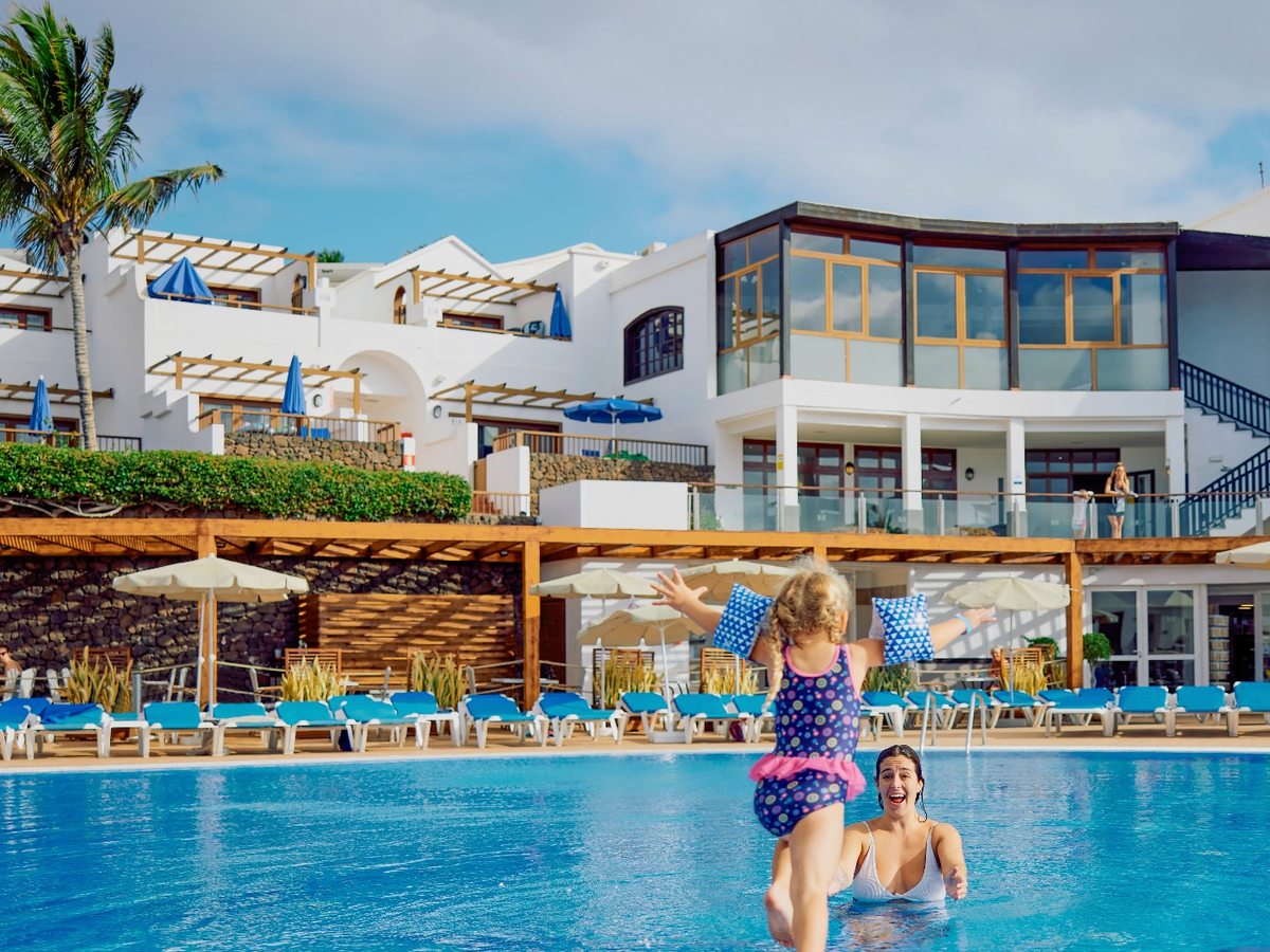 A woman and a young girl in armbands playing in a large outdoor pool, with rows of blue sunloungers, parasols and the resort's white two-storey blocks behind.