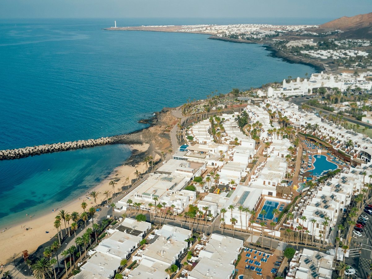 An aerial view along the coast at Playa Blanca, with a sandy bay and rock breakwater on the left and the resort's white low-rise blocks, pools and palms spread along the shore.