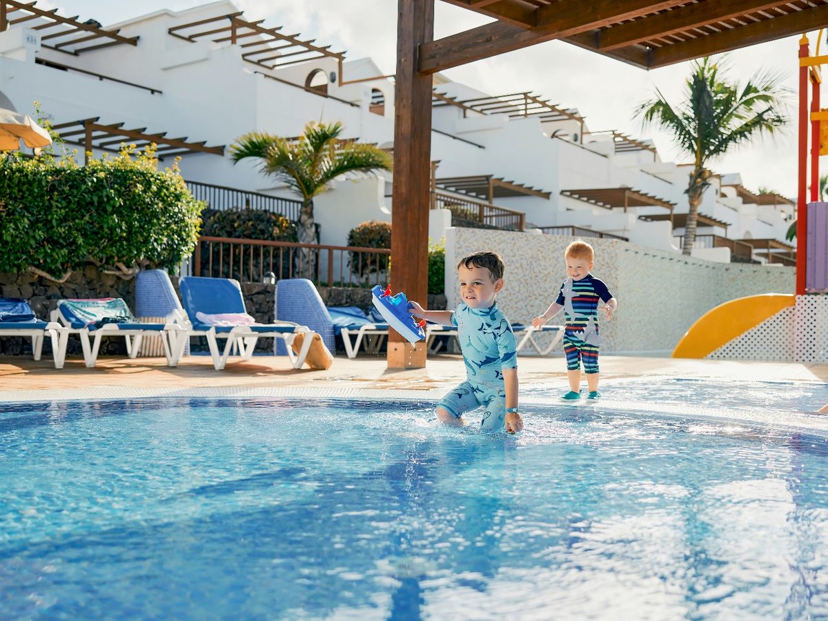 Two toddlers in swimsuits playing at the edge of a shallow pool, with a small yellow slide to the right and the resort's white terraced apartments behind.