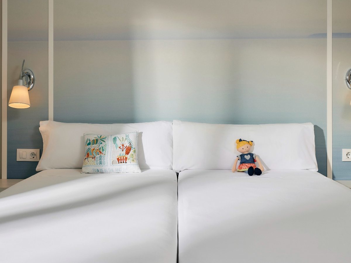 Two single beds pushed together and made up in white, with a pale blue panelled headboard, a children's cushion and a rag doll on the pillows.