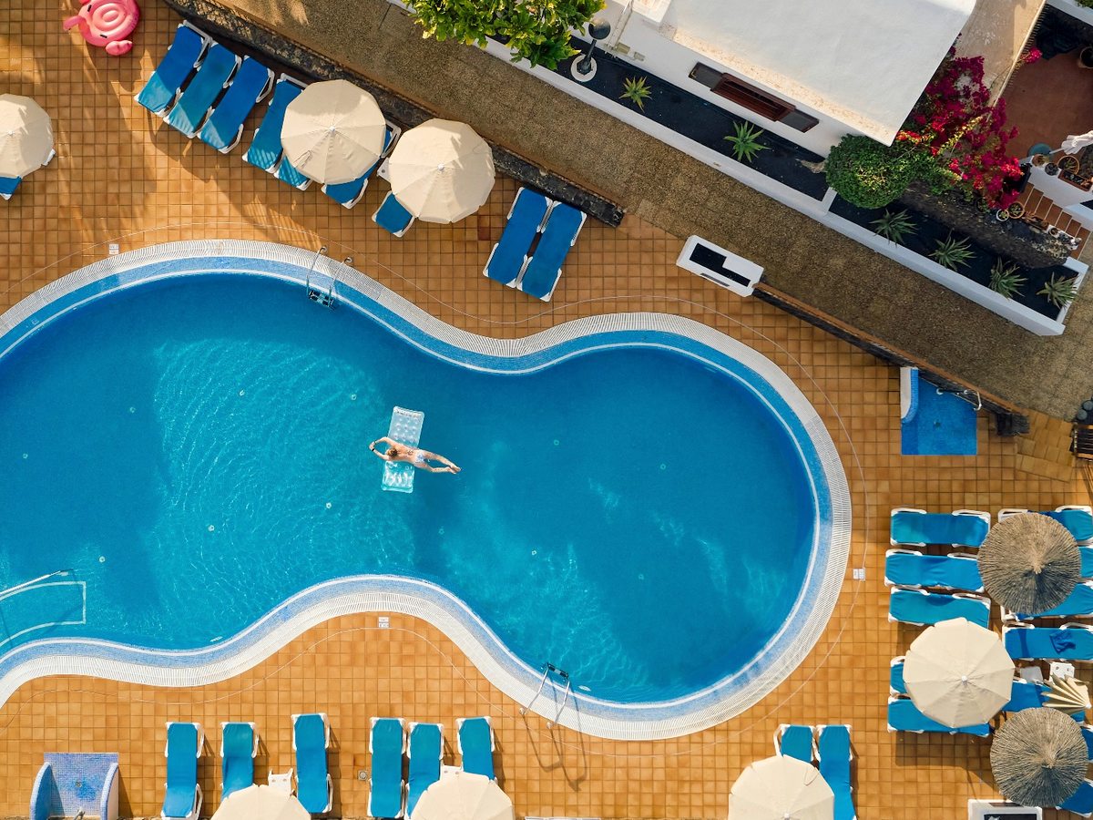 An overhead view of a curved pool edged with terracotta tiling, rows of blue sunloungers and cream parasols around it and one swimmer on a lilo.